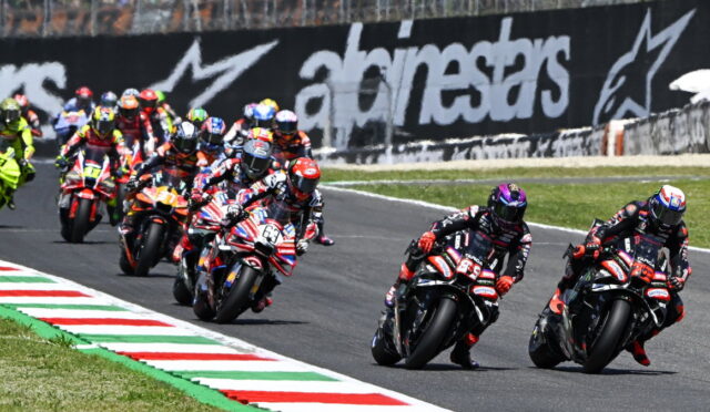 Motorcycle race with riders leaning into a curve on a track, colorful helmets and sponsor logos in view.