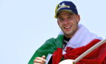 Smiling man wearing a blue Michelin cap with an Italian flag draped over his shoulders, holding a flagpole against a clear sky.