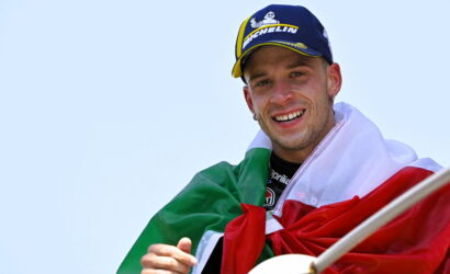 Smiling man wearing a blue Michelin cap with an Italian flag draped over his shoulders, holding a flagpole against a clear sky.