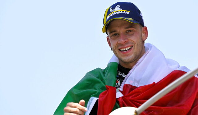 Smiling man wearing a blue Michelin cap with an Italian flag draped over his shoulders, holding a flagpole against a clear sky.