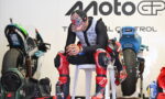 MotoGP rider in red-and-black protective gear sits on a white chair in a workshop, with bikes and gear in the background.