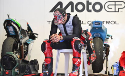 MotoGP rider in red-and-black protective gear sits on a white chair in a workshop, with bikes and gear in the background.