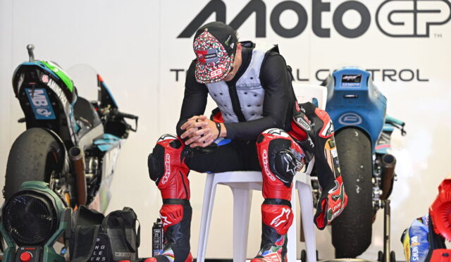 MotoGP rider in red-and-black protective gear sits on a white chair in a workshop, with bikes and gear in the background.