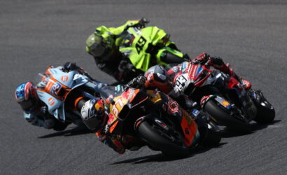 Group of sport motorcycles leaning into a corner at a racetrack, riders in colorful helmets and suits close together in a tight pack.