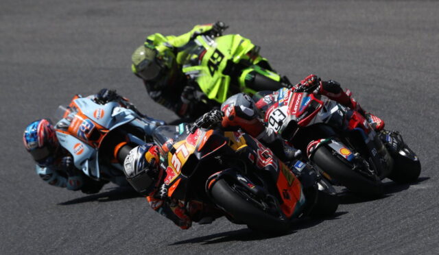 Group of sport motorcycles leaning into a corner at a racetrack, riders in colorful helmets and suits close together in a tight pack.