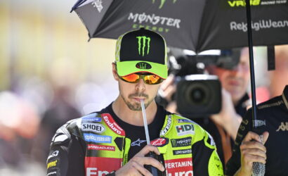 MotoGP rider in a neon green and black racing suit drinks through a straw under a Monster Energy umbrella, with a camera crew in the background.
