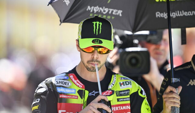 MotoGP rider in a neon green and black racing suit drinks through a straw under a Monster Energy umbrella, with a camera crew in the background.