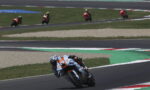 Motorcycle racer in white-orange suit leaning into a curve on a race track, with other riders in the distance behind on the straights.