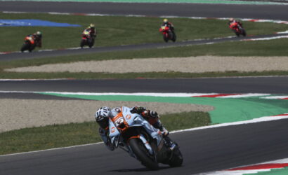 Motorcycle racer in white-orange suit leaning into a curve on a race track, with other riders in the distance behind on the straights.