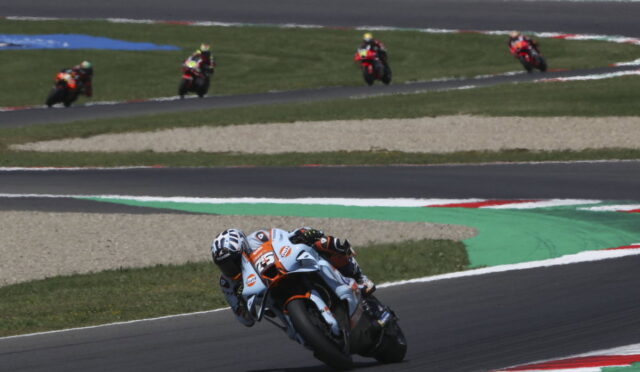 Motorcycle racer in white-orange suit leaning into a curve on a race track, with other riders in the distance behind on the straights.