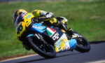Motorcycle racer in a yellow-and-blue suit leaning into a turn on a blue racing bike with number 46 and sponsor logos visible on the fairing.