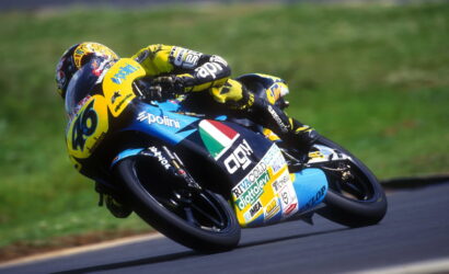 Motorcycle racer in a yellow-and-blue suit leaning into a turn on a blue racing bike with number 46 and sponsor logos visible on the fairing.