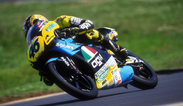 Motorcycle racer in a yellow-and-blue suit leaning into a turn on a blue racing bike with number 46 and sponsor logos visible on the fairing.
