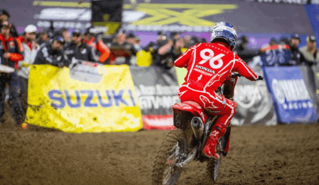 Motocross rider in red gear, number 96, speeding through a muddy track with a crowd and sponsor banners in the background.