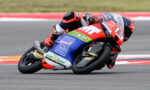 Motorcycle racer in red-and-blue suit leaning into a turn on a race track with sponsor decals visible on the bike.