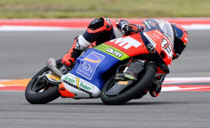 Motorcycle racer in red-and-blue suit leaning into a turn on a race track with sponsor decals visible on the bike.