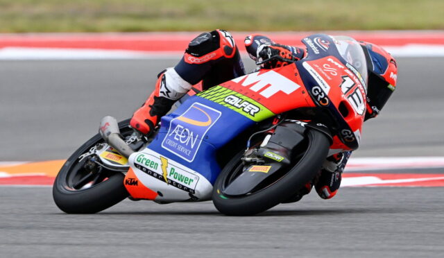 Motorcycle racer in red-and-blue suit leaning into a turn on a race track with sponsor decals visible on the bike.