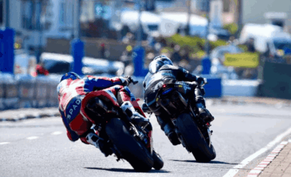 Two motorcycle racers lean into a sharp turn on an urban street race, side by side in full gear and bright leathers.