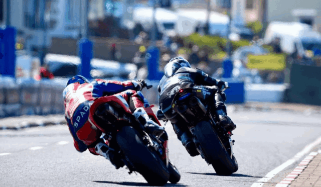 Two motorcycle racers lean into a sharp turn on an urban street race, side by side in full gear and bright leathers.