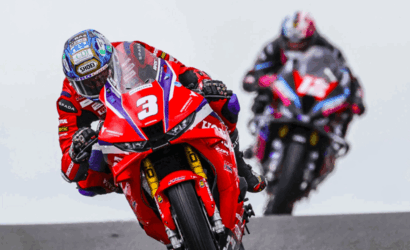 Two motorcycle racers lean into a turn on a race track; the rider in front wears red with the number 3 on the bike.
