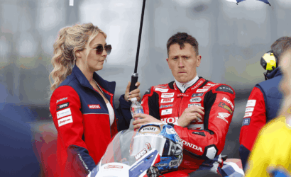 Moto racer in a red Honda racing suit sits with helmet on his lap while a woman in team gear holds a large umbrella over him.