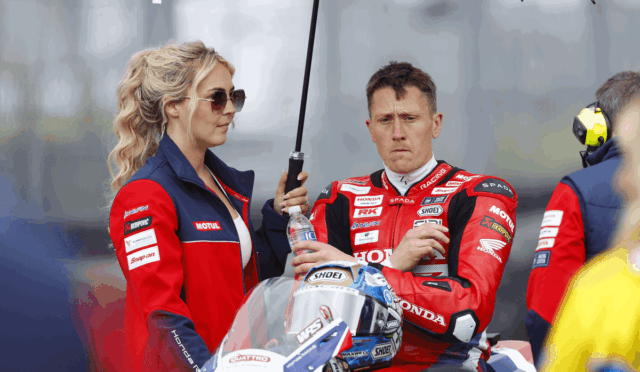 Moto racer in a red Honda racing suit sits with helmet on his lap while a woman in team gear holds a large umbrella over him.