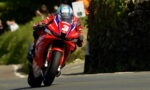 Motorcycle racer in red livery leans into a turn on a red sport bike during a race, helmeted and focused, on a winding road.