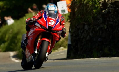 Motorcycle racer in red livery leans into a turn on a red sport bike during a race, helmeted and focused, on a winding road.
