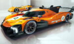 Orange Le Mans–style race car with number 4 and sponsor decals, parked in a white showroom with a second orange car in the background.