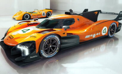 Orange Le Mans–style race car with number 4 and sponsor decals, parked in a white showroom with a second orange car in the background.