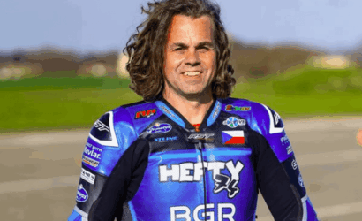 Male motorcycle racer in a blue racing suit, smiling, standing outdoors with a blurred green field background.