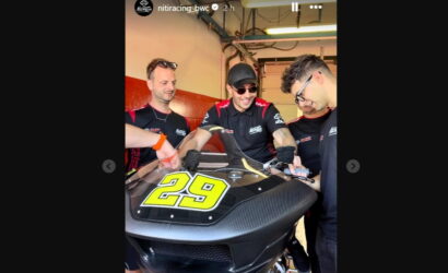 Team members in matching black and red shirts work on a race bike with the number 29 in a garage.