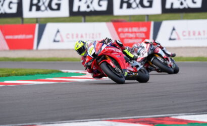 Two motorcycle racers lean into a tight turn on a race track, red bikes close together with neon-yellow helmets visible under a banners-lined barrier.