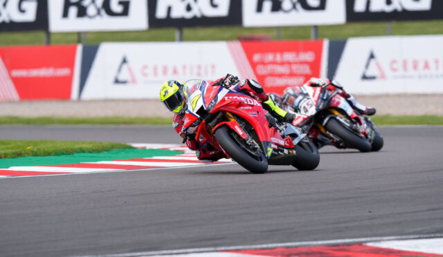 Two motorcycle racers lean into a tight turn on a race track, red bikes close together with neon-yellow helmets visible under a banners-lined barrier.