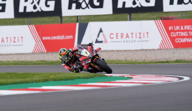 Motorcycle racer leans into a tight left-hand turn on a race track, red‑white curbs visible ahead.