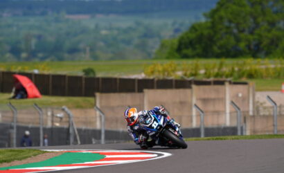 Motorcycle racer in blue and white riding gear leaning into a sharp turn on a race track, bike numbered 19 with a blurred grassy backdrop and red-green-white curb in the foreground.