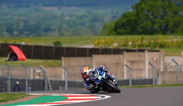 Motorcycle racer in blue and white riding gear leaning into a sharp turn on a race track, bike numbered 19 with a blurred grassy backdrop and red-green-white curb in the foreground.
