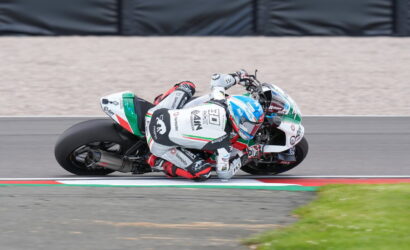 Motorcycle racer in white suit leaning sharply into a turn on a race track, blue and white bike with red accents.