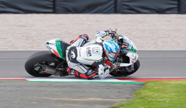 Motorcycle racer in white suit leaning sharply into a turn on a race track, blue and white bike with red accents.