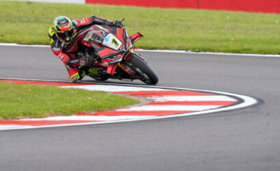Motorcycle racer in red-black gear leaning into a sharp turn on a race track with red-and-white curbs and green grass nearby