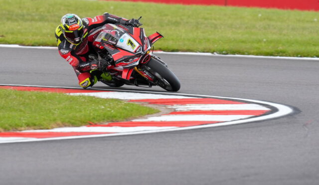 Motorcycle racer in red-black gear leaning into a sharp turn on a race track with red-and-white curbs and green grass nearby