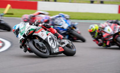 Motorcycle race on a track with several riders leaning into a sharp turn; foreground rider wears white, green, and red livery and blue helmet.",
