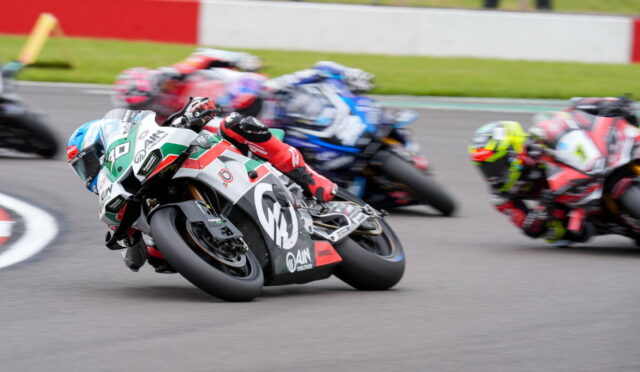 Motorcycle race on a track with several riders leaning into a sharp turn; foreground rider wears white, green, and red livery and blue helmet.",