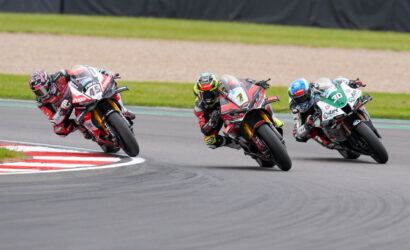Three motorcycle racers lean into a left-hand turn on a race track, wearing colorful leathers and helmets.