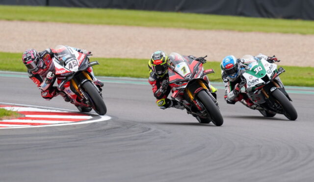 Three motorcycle racers lean into a left-hand turn on a race track, wearing colorful leathers and helmets.