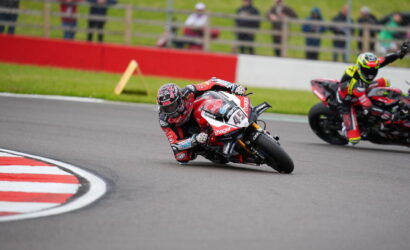 Motorcycle racer in red-and-white gear leans into a left-hand turn on a race track, with a second rider behind and spectators in the background.