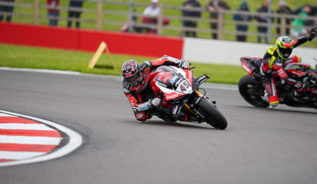 Motorcycle racer in red-and-white gear leans into a left-hand turn on a race track, with a second rider behind and spectators in the background.
