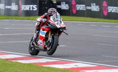 Motorcycle racer in red and white gear leans into a high-speed corner on a race track with barriers in the background.