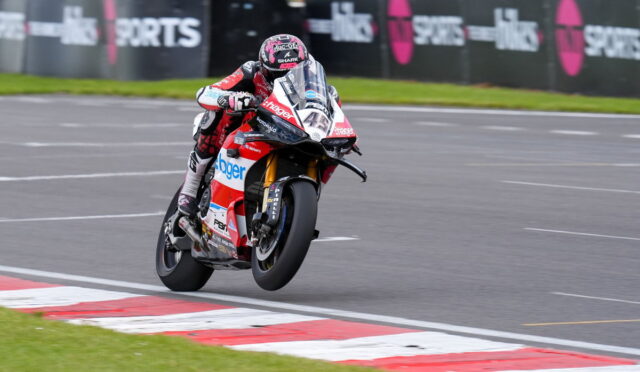 Motorcycle racer in red and white gear leans into a high-speed corner on a race track with barriers in the background.