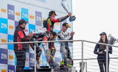 Riders on a podium celebrate with trophies as a woman in a cap holds a large trophy to the right, banners and logos in the background.